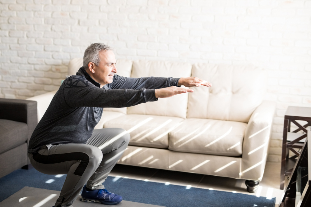 SeniorManDoingSquatsAndExercisingInLivingRoomAt - welltica.dk - Vejen til et bedre liv