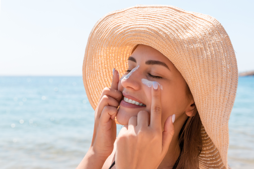 SmilingWomanInHatIsApplyingSunscreenOnHerFace - welltica.dk - Vejen til et bedre liv