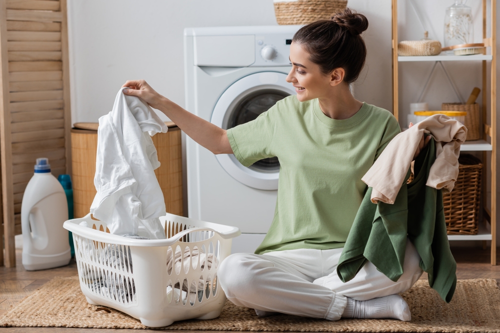 HappyYoungWomanSortingClothesNearWashingMachineInLaundry - welltica.dk - Vejen til et bedre liv