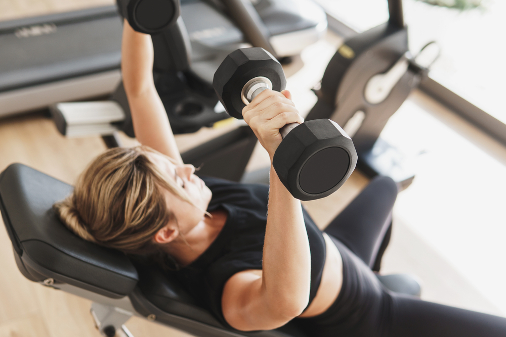YoungAthleticWomanDoingBenchPressUsingDumbbellsInGym - welltica.dk - Vejen til et bedre liv