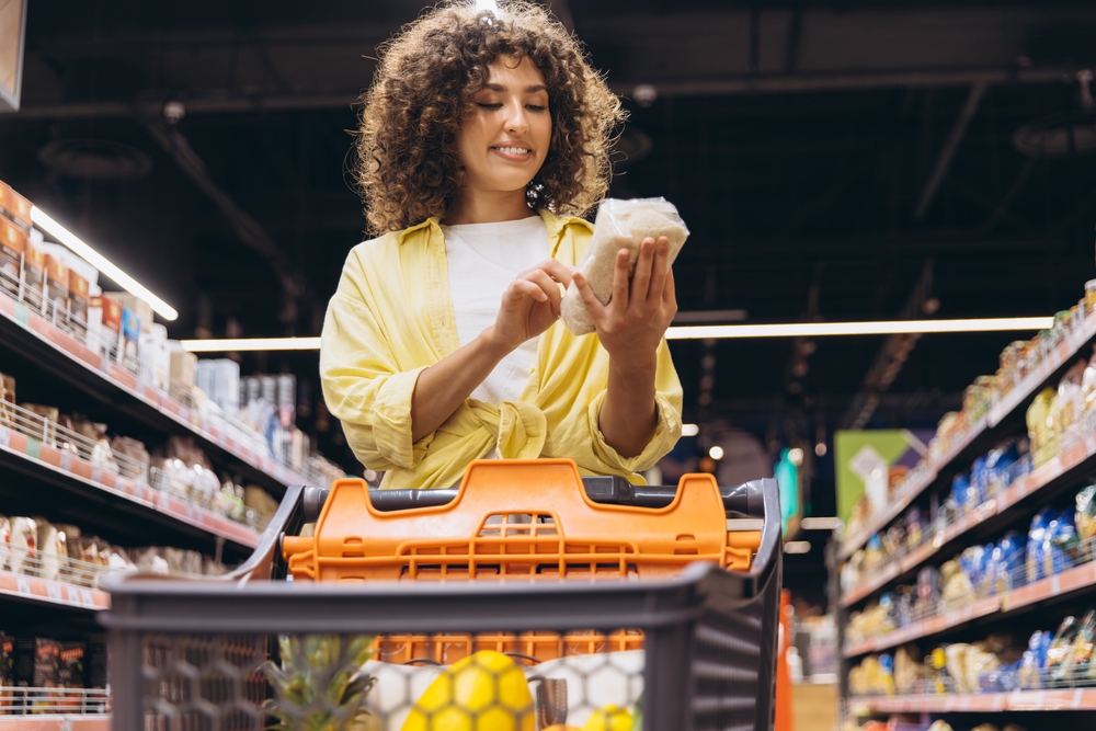 SmilingCurlyWomanPushingShoppingCartChoosingFoodProductsReading - welltica.dk - Vejen til et bedre liv